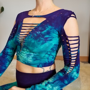 Pu&ograve; includere: Un top corto a maniche lunghe con stampa tie-dye, un profondo scollo a V e dettagli tagliati sulle maniche. Il top &egrave; in tonalit&agrave; di blu e verde.