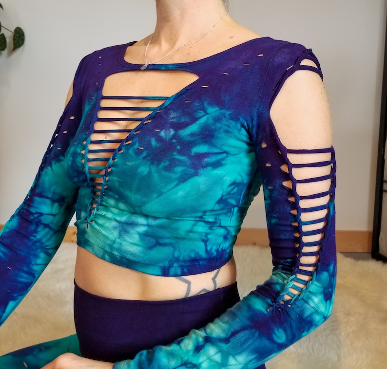 Pu&ograve; includere: Un top corto a maniche lunghe con stampa tie-dye, un profondo scollo a V e dettagli tagliati sulle maniche. Il top &egrave; in tonalit&agrave; di blu e verde.