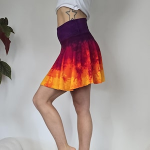 May include: A tie-dye mini skirt with a wide purple waistband. The skirt transitions from red to orange and yellow. The skirt is flared and has a soft, flowing appearance. The garment is made of a lightweight fabric.