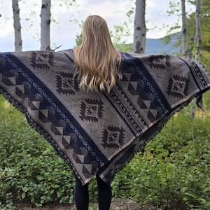 Op de afbeelding: Een vrouw draagt een bruin en blauw gepatroneerd poncho met franjes. De poncho heeft een geometrisch ontwerp en is gemaakt van een zachte, geweven stof.