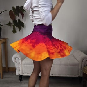 May include: A swirling skirt in shades of orange, red, and purple. The skirt is flared and appears to be made of a lightweight fabric. The person is wearing a white hooded sweatshirt.