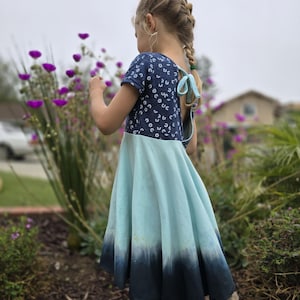 May include: A young girl wearing a blue and white patterned dress with a light blue and dark blue ombre skirt. The dress has a tie at the back. The girl is standing in a garden with purple flowers.