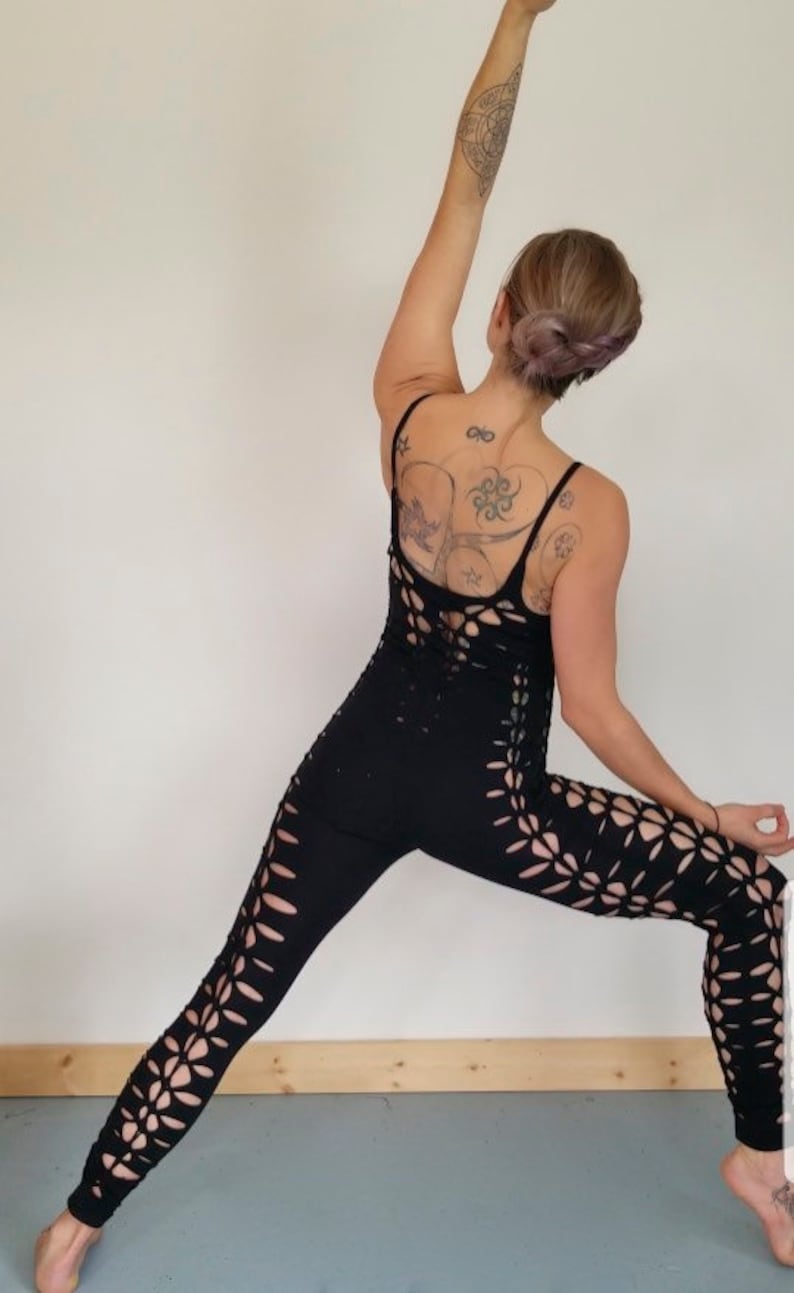 May include: A woman wearing a black, cut-out jumpsuit with a floral design. She is standing in a yoga pose with her arm raised above her head.