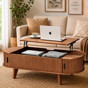 May include: A wooden coffee table with a lift-top mechanism, featuring a fluted exterior design and rounded ends. The table has an open storage compartment containing two folded grey blankets, with a silver laptop and a white mug on top.