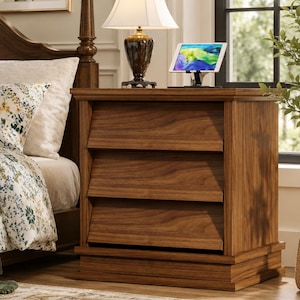 May include: A wooden nightstand featuring three horizontal drawers with angled fronts and a rectangular base. The piece has a warm brown wood grain finish and is positioned next to a bed, supporting a table lamp and a tablet on a stand.