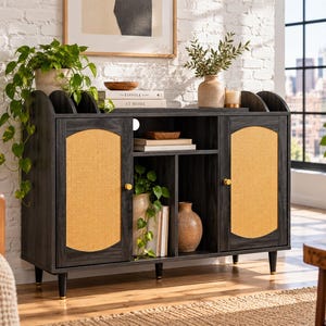 May include: A dark charcoal wood sideboard featuring two cabinet doors with arched light-tan cane webbing panels and gold-toned knobs. The unit has an open center shelf, a flat top surface, and four tapered legs with gold-toned feet.