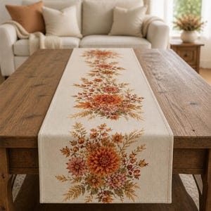 Könnte beinhalten: Ein rechteckiger Tischläufer aus hellbeigem Stoff mit einem Druckmotiv aus herbstlichen Blumen, darunter große orangefarbene Dahlien, kleinere orange-rosa Blüten sowie verschiedenes grünes und braunes Blattwerk in der Mitte.
