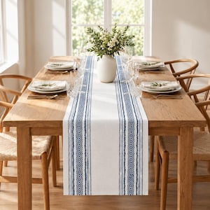 Op de afbeelding: Een lange rechthoekige tafelloper met een wit midden en twee verticale marineblauwe strepen met geometrische ruitpatronen en gestructureerde lijnen. De loper ligt op een houten eettafel met witte borden en glazen.
