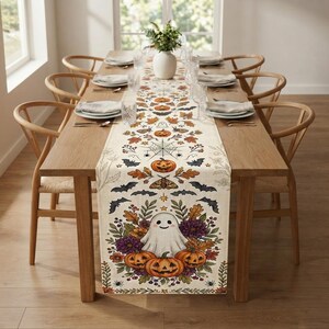 May include: A long rectangular table runner featuring a cream-coloured fabric printed with a repeating Halloween pattern of orange pumpkins, black bats, moths, spiderwebs, autumn leaves, and a central illustration of a white ghost among flowers.
