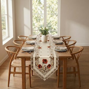 May include: A rectangular wooden dining table with eight light wood chairs featuring woven seats. A cream-colored table runner with embroidered red poppy flowers and scalloped edges runs down the center, topped with a white vase of greenery.