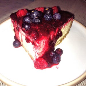Pode incluir: Uma fatia triangular de cheesecake num prato branco, coberta com uma compota espessa de frutos vermelhos, mirtilos frescos e framboesas. O recheio branco cremoso assenta numa base dourada, com o molho de fruta a escorrer pelos lados.