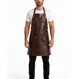 May include: A dark brown apron featuring a textured crocodile skin pattern. The apron includes a chest pocket with a metal D-ring, two large lower pockets, and adjustable shoulder straps secured with metal hardware and rivets for a structured fit.