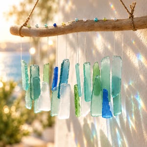 May include: A hanging mobile featuring multiple translucent sea glass pieces in shades of blue, green, and white, suspended by thin strings from a natural driftwood branch. Small colourful beads are attached along the top of the wood.