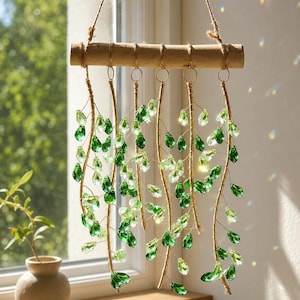 Può includere: Una decorazione da parete sospesa composta da un ramo di legno orizzontale con sei fili di metallo dorato. Ogni filo è decorato con molteplici cristalli di vetro a forma di goccia in varie tonalità di verde chiaro e verde scuro.