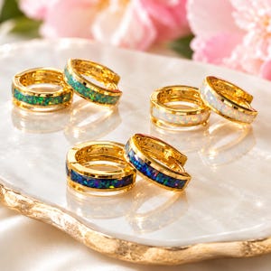 May include: Three pairs of gold hoop earrings featuring inlaid stone bands. One pair has green iridescent inlays, one pair has white iridescent inlays, and one pair has dark blue iridescent inlays. Each hoop has a hinged closure mechanism.