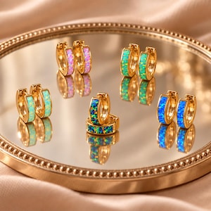 May include: A collection of five pairs of gold hoop earrings, each featuring a different colored inlay. The colors include pink, light green, mint green, dark blue, and a multi-colored dark blue, all with a shimmering, textured finish.