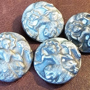 May include: Four circular silver-toned metal buttons featuring a raised relief design of a figure holding a fan. Each button displays a detailed embossed pattern with a weathered, metallic finish and a rounded edge, presented as a set of matching fasteners.