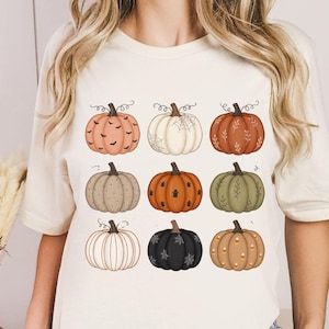 Könnte beinhalten: Ein cremefarbenes Kurzarm-T-Shirt mit einem Grafikdruck von neun stilisierten Kürbissen, die in einem Raster angeordnet sind. Die Kürbisse variieren in der Farbe, darunter Orange-, Weiß-, Grün- und Schwarztöne, jeweils verziert mit Mustern wie Blättern und Punkten.