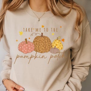 Könnte beinhalten: Ein beigefarbenes Sweatshirt mit Rundhalsausschnitt und einem Grafikdruck von vier Kürbissen mit verschiedenen Mustern wie Punkten, Leopardenmuster und Sternen. Der Text "TAKE ME TO THE pumpkin patch" ist in Braun um die Kürbisse gedruckt.