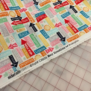 May include: White fabric with a repeating pattern of colorful city names and arrows. The fabric has the text "michael miller FABRICS Patt# CX8436 Many Directions #makingitfun" printed on the bottom edge.