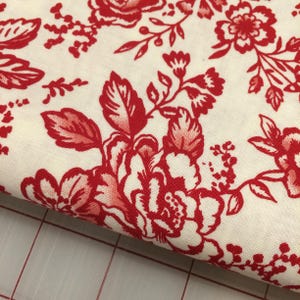 May include: Close-up of a fabric with a cream background and a repeating pattern of red floral designs. The flowers and leaves are detailed with varying shades of red, creating a classic, vintage aesthetic. The fabric is likely used for sewing or crafting.