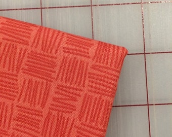 May include: A close-up of a piece of coral orange fabric with a woven pattern of thin red lines. The fabric is lying on a white surface with a grid pattern.