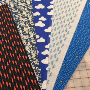 May include: Assortment of fabric swatches with various patterns. Designs include orange carrots on navy, birds on white and navy, white clouds on blue, seahorses on white, and blue fish on blue. The fabrics are arranged side by side, showcasing the different patterns.