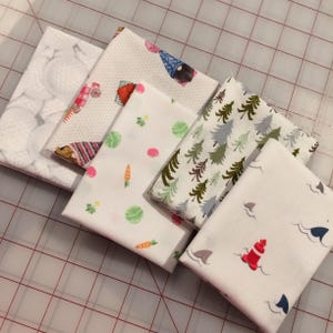 May include: A collection of folded fabric squares with various patterns. Designs include white with gray circles, gnomes, vegetables, evergreen trees, and a nautical theme with a lighthouse and sharks. The fabrics are arranged on a grid.