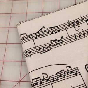 May include: A close-up of white fabric with a black musical note pattern. The fabric has a subtle texture and is perfect for crafting projects.