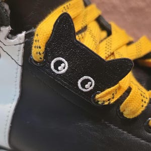 Könnte beinhalten: Ein schwarzer, katzenförmiger Schnürsenkel-Anhänger an einem schwarzen Schuh mit leuchtend gelben Schnürsenkeln. Der Anhänger hat gestickte weiße, kreisförmige Augen und spitze Ohren und ist zwischen den Ösen platziert, durch die die gelben Schnürsenkel verlaufen.
