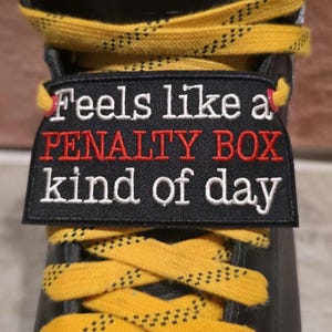 May include: A black ice skate featuring bright yellow laces with a black speckled pattern. A rectangular black embroidered patch is attached to the laces, displaying the white and red text: Feels like a PENALTY BOX kind of day.