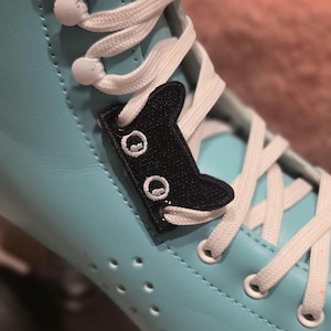 May include: A light blue roller skate featuring white laces threaded through a black embroidered cat-shaped lace charm. The charm has two white circular eyes and is positioned on the front of the skate, secured by the white shoelaces.