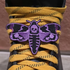 May include: A black leather boot featuring bright yellow laces threaded through a purple and black embroidered moth patch. The moth design includes a skull detail on its thorax and intricate wing patterns, secured directly onto the laces.