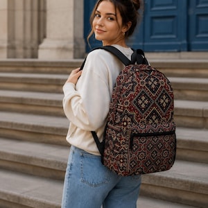 May include: A backpack featuring a dark-toned fabric with a repeating geometric pattern in shades of deep red, navy blue, and cream. The bag includes a main zippered compartment, a smaller front zippered pocket, and adjustable black shoulder straps.