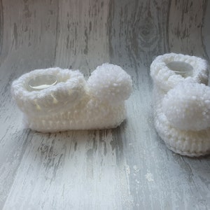 May include: Two white crocheted baby booties with pom-poms. The booties are made with a soft, fluffy yarn and have a simple, classic design.