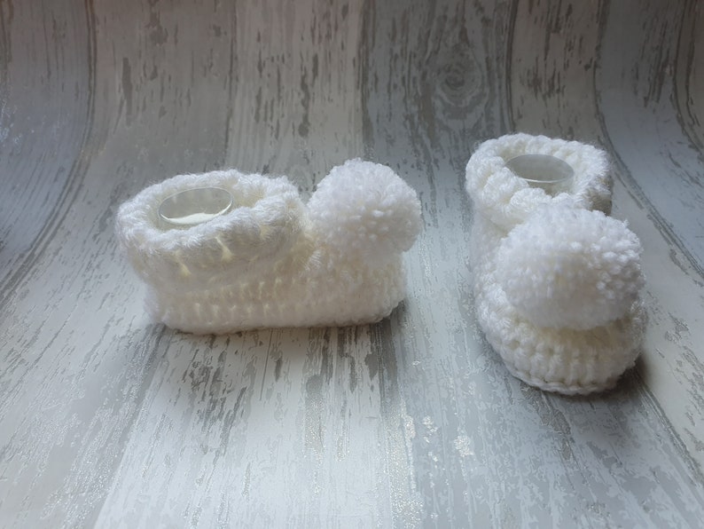 May include: Two white crocheted baby booties with pom-poms. The booties are made with a soft, fluffy yarn and have a simple, classic design.