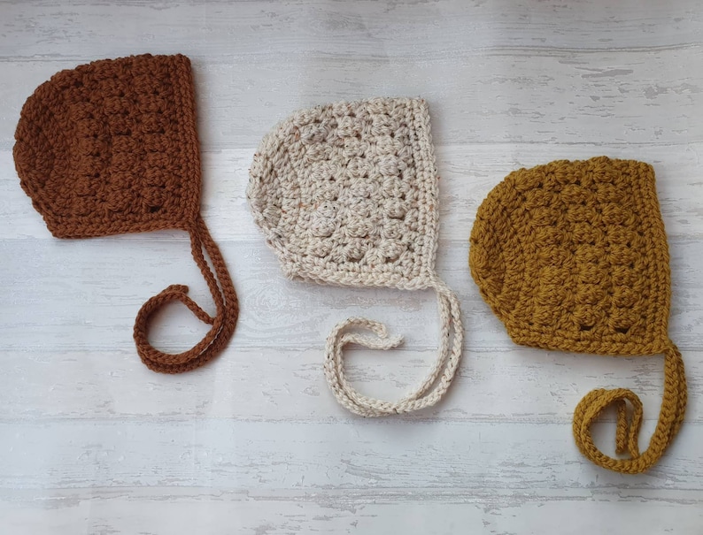 K&ouml;nnte beinhalten: Drei geh&auml;kelte Babyhauben in Braun, Creme und Senfgelb. Jede Haube hat eine lange Schleife, die unter dem Kinn gebunden werden kann.