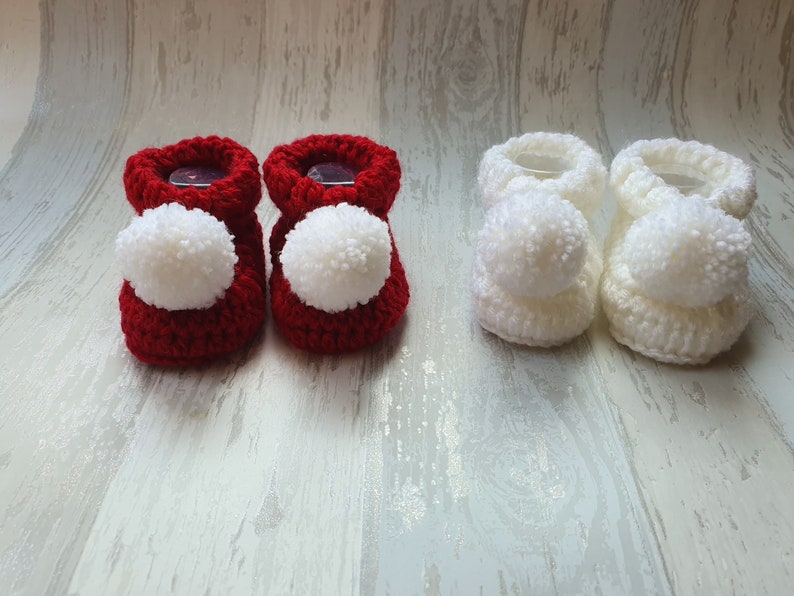 May include: Two pairs of crocheted baby booties with pom poms. One pair is red and the other is white.