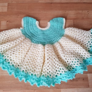K&ouml;nnte beinhalten: Ein geh&auml;keltes Babykleid in Wei&szlig; und T&uuml;rkis. Das Kleid hat einen R&uuml;schenrock und einen runden Ausschnitt.