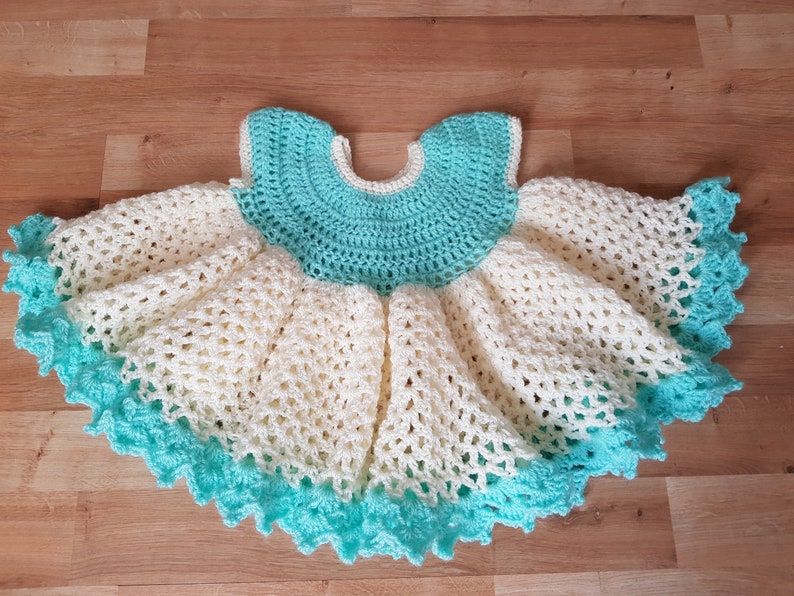 K&ouml;nnte beinhalten: Ein geh&auml;keltes Babykleid in Wei&szlig; und T&uuml;rkis. Das Kleid hat einen R&uuml;schenrock und einen runden Ausschnitt.