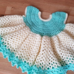 K&ouml;nnte beinhalten: Ein geh&auml;keltes Babykleid in Wei&szlig; und T&uuml;rkis. Das Kleid hat einen Volantensaum und einen runden Ausschnitt.