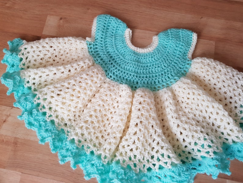 K&ouml;nnte beinhalten: Ein geh&auml;keltes Babykleid in Wei&szlig; und T&uuml;rkis. Das Kleid hat einen Volantensaum und einen runden Ausschnitt.