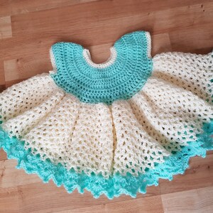 K&ouml;nnte beinhalten: Ein geh&auml;keltes Babykleid in Wei&szlig; und T&uuml;rkis. Das Kleid hat einen gewellten Saum und einen runden Ausschnitt.