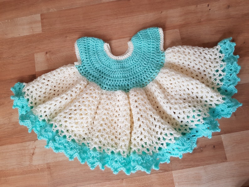 K&ouml;nnte beinhalten: Ein geh&auml;keltes Babykleid in Wei&szlig; und T&uuml;rkis. Das Kleid hat einen gewellten Saum und einen runden Ausschnitt.