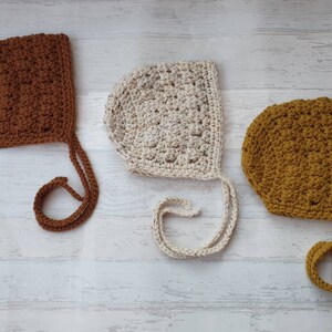K&ouml;nnte beinhalten: Drei geh&auml;kelte Babyhauben in Braun, Wei&szlig; und Senfgelb mit B&auml;ndern.
