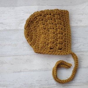 K&ouml;nnte beinhalten: Eine geh&auml;kelte Babyhaube in senfgelber Farbe mit einer langen Schleife.