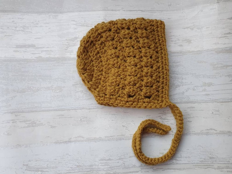 K&ouml;nnte beinhalten: Eine geh&auml;kelte Babyhaube in senfgelber Farbe mit einer langen Schleife.