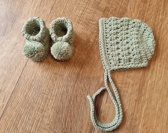 May include: A sage green crochet baby set, including a pair of booties with pom-poms and a bonnet with ties. The handmade set is displayed on a wooden surface. The booties and bonnet are made of soft yarn.