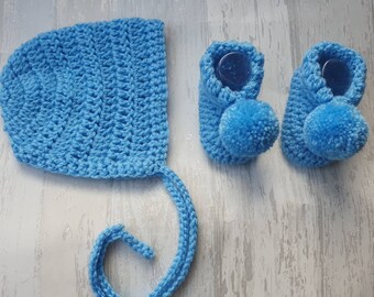 May include: A set of baby booties and a bonnet in a light blue color. The booties have a pom-pom on each toe and the bonnet has a long tie.