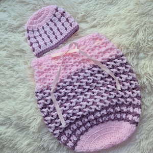 May include: A handmade crochet baby cocoon and matching hat set. The cocoon is a soft pink and purple, with a textured stitch pattern. A light pink ribbon is tied at the top. The hat is a matching pink and purple color.
